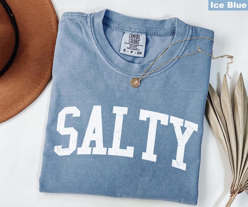 Puede incluir: Una camiseta azul claro con la palabra "SALTY" impresa en blanco en la parte delantera. La camiseta est&aacute; hecha de una tela suave y c&oacute;moda y tiene un corte holgado.