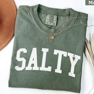 Puede incluir: Una camiseta verde con la palabra "SALTY" impresa en blanco en la parte delantera. La camiseta est&aacute; hecha de una tela suave y c&oacute;moda y tiene un corte holgado.