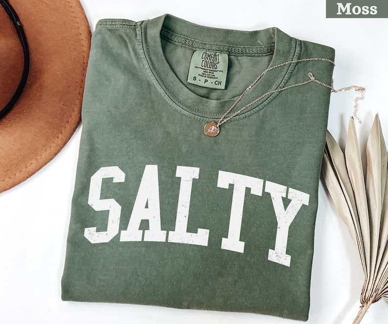 Puede incluir: Una camiseta verde con la palabra "SALTY" impresa en blanco en la parte delantera. La camiseta est&aacute; hecha de una tela suave y c&oacute;moda y tiene un corte holgado.