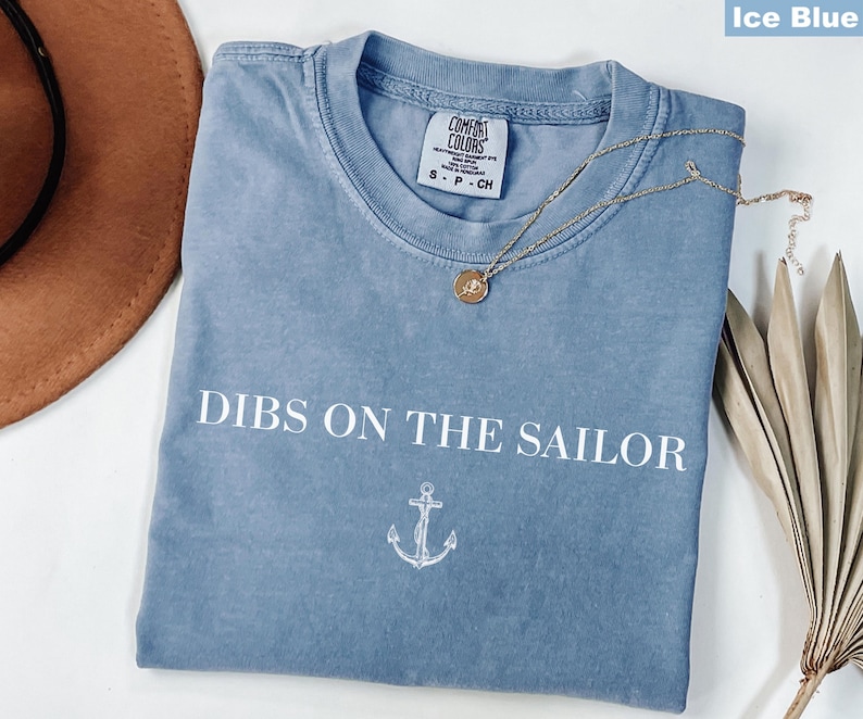 May include: Ice blue t-shirt with the text "DIBS ON THE SAILOR" and an anchor graphic. The shirt has a crew neck and short sleeves. A gold necklace with a pendant is draped across the shirt.