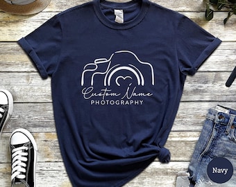 May include: Navy blue t-shirt with a white graphic of a camera and the words "Custom Name Photography". The shirt is made of a soft material and has rolled-up sleeves. A pair of black and white sneakers and blue jeans are visible.