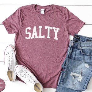 Puede incluir: Camiseta de color marr&oacute;n heather con la palabra "SALTY" impresa en blanco en la parte delantera. La camiseta est&aacute; sobre una superficie blanca con un par de zapatillas blancas y unos vaqueros azules.