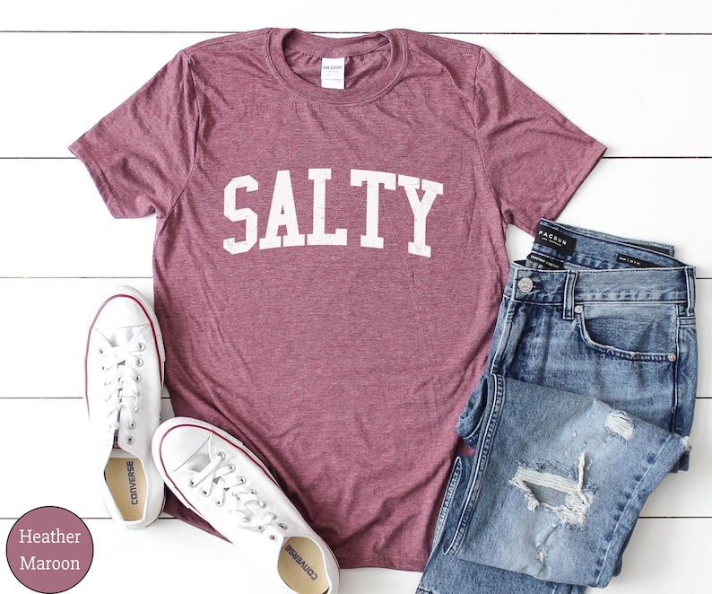 Puede incluir: Camiseta de color marr&oacute;n heather con la palabra "SALTY" impresa en blanco en la parte delantera. La camiseta est&aacute; sobre una superficie blanca con un par de zapatillas blancas y unos vaqueros azules.