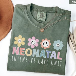May include: Moss green t-shirt with a floral design and the words "NEONATAL INTENSIVE CARE UNIT" in pastel colors. The shirt has a crew neck and short sleeves. A gold necklace with a pendant is draped across the shirt.