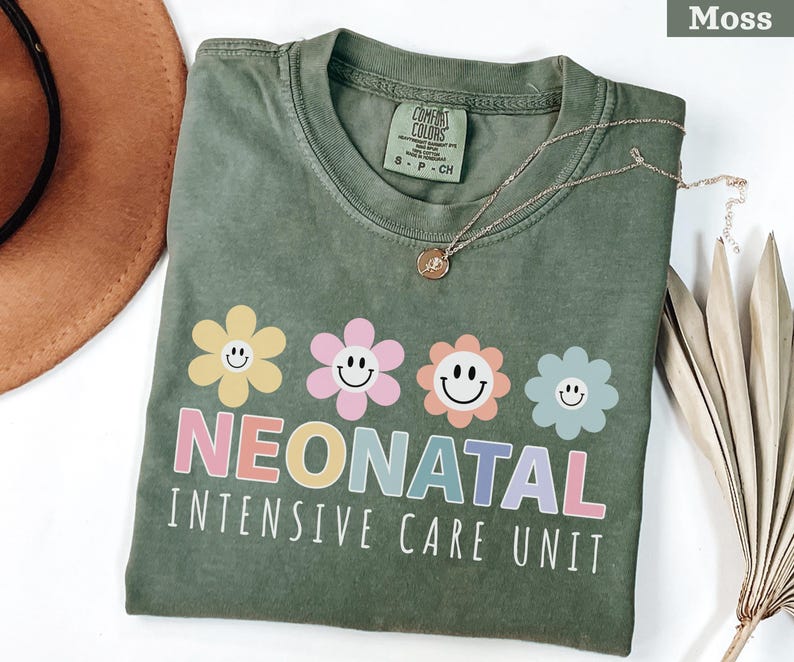 May include: Moss green t-shirt with a floral design and the words "NEONATAL INTENSIVE CARE UNIT" in pastel colors. The shirt has a crew neck and short sleeves. A gold necklace with a pendant is draped across the shirt.