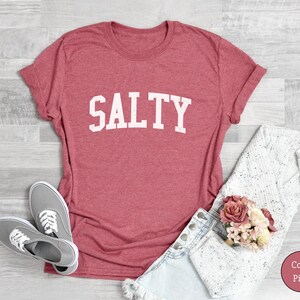 Puede incluir: Una camiseta de color coral con la palabra "SALTY" impresa en blanco en la parte delantera. La camiseta est&aacute; hecha de un material suave y c&oacute;modo y tiene un corte holgado.