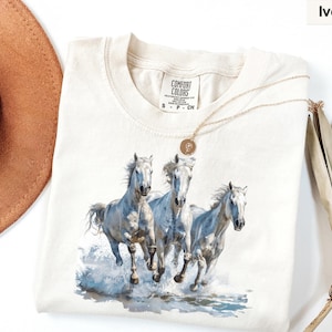 May include: Ivory-colored Comfort Colors t-shirt featuring a watercolor illustration of three white horses running through water. The horses have flowing manes and tails, with water splashing around their hooves. A gold necklace is draped over the shirt.