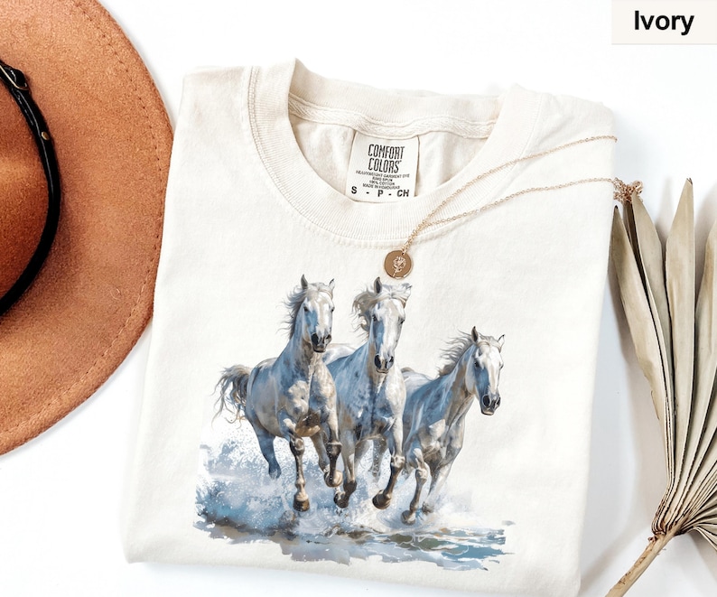 May include: Ivory-colored Comfort Colors t-shirt featuring a watercolor illustration of three white horses running through water. The horses have flowing manes and tails, with water splashing around their hooves. A gold necklace is draped over the shirt.
