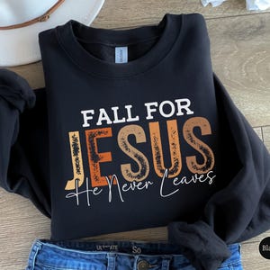 K&ouml;nnte beinhalten: Schwarzes Sweatshirt mit dem Text "FALL FOR JESUS He Never Leaves" in Wei&szlig;, Orange und Braun. Das Sweatshirt ist auf einer blauen Jeans gefaltet. Ein wei&szlig;er Hut ist im Hintergrund zu sehen.