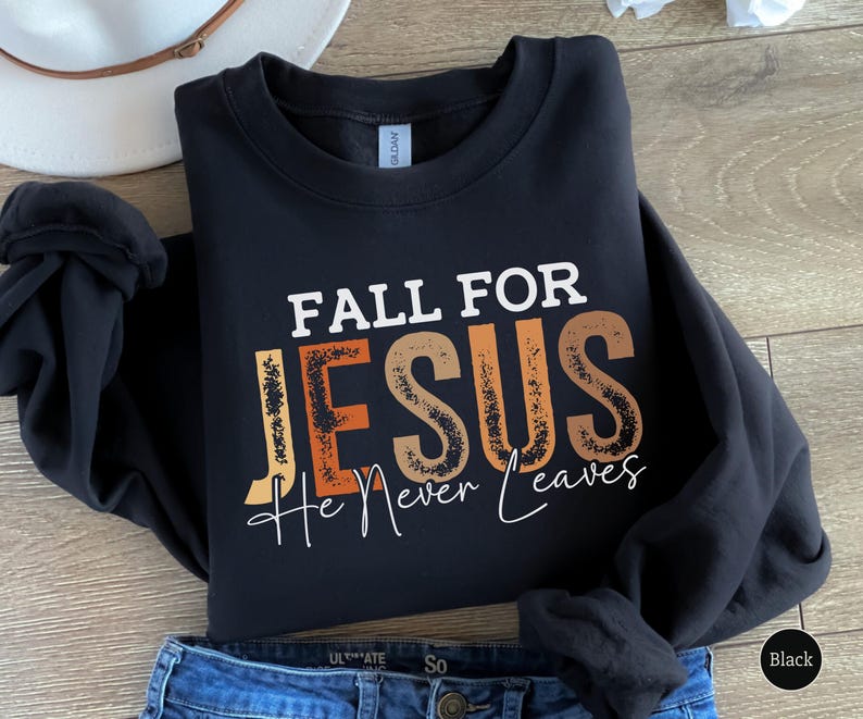 K&ouml;nnte beinhalten: Schwarzes Sweatshirt mit dem Text "FALL FOR JESUS He Never Leaves" in Wei&szlig;, Orange und Braun. Das Sweatshirt ist auf einer blauen Jeans gefaltet. Ein wei&szlig;er Hut ist im Hintergrund zu sehen.