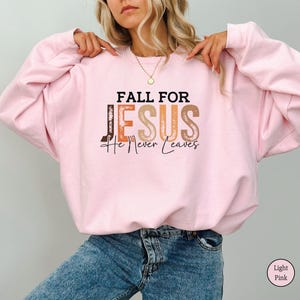 K&ouml;nnte beinhalten: Hellrosa Sweatshirt mit dem Text "FALL FOR JESUS He Never Leaves" in braunen, orangefarbenen und beigen Buchstaben. Das Sweatshirt hat eine einfarbige hellrosa Farbe.