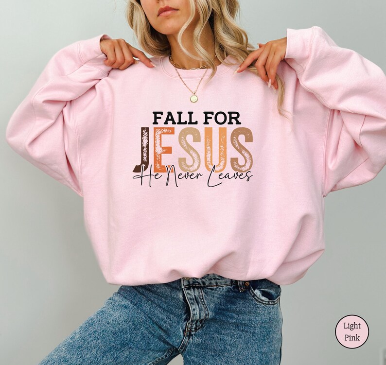 K&ouml;nnte beinhalten: Hellrosa Sweatshirt mit dem Text "FALL FOR JESUS He Never Leaves" in braunen, orangefarbenen und beigen Buchstaben. Das Sweatshirt hat eine einfarbige hellrosa Farbe.