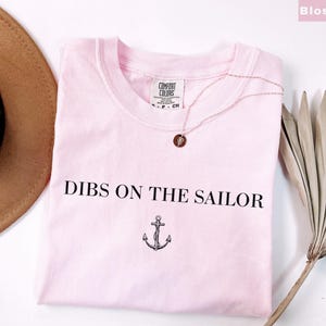 May include: A light pink t-shirt with the text "DIBS ON THE SAILOR" and an anchor graphic. A gold-colored necklace with a pendant rests on the shirt. A brown hat and dried palm frond are in the background.