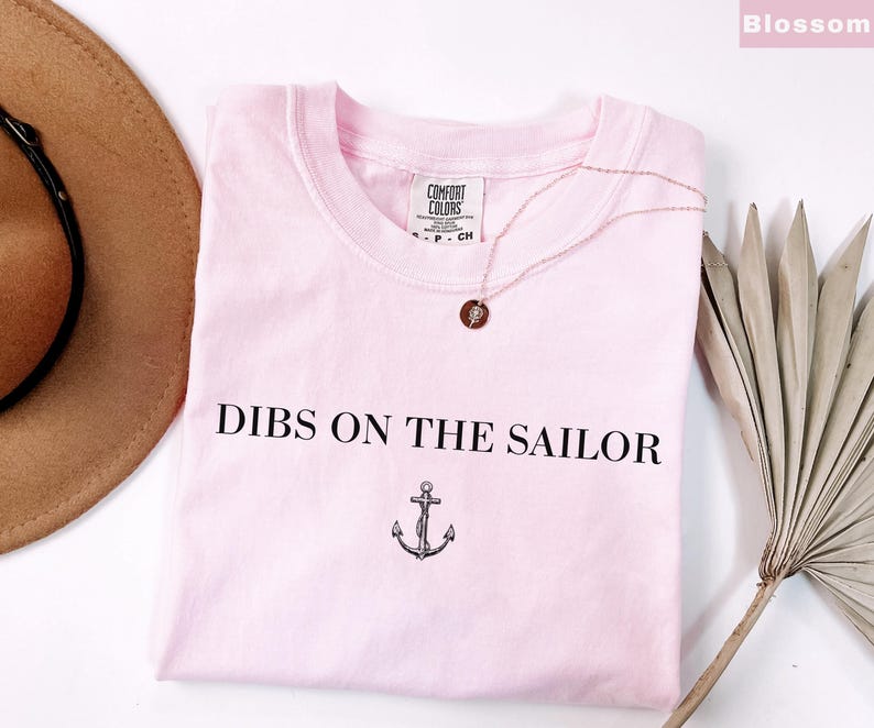 May include: A light pink t-shirt with the text "DIBS ON THE SAILOR" and an anchor graphic. A gold-colored necklace with a pendant rests on the shirt. A brown hat and dried palm frond are in the background.