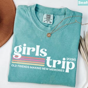 May include: Seafoam green t-shirt with the words "girls trip 2026" in white, with a colorful striped design. The shirt also has the text "OLD FRIENDS MAKING NEW MEMORIES". A gold necklace is draped across the shirt.