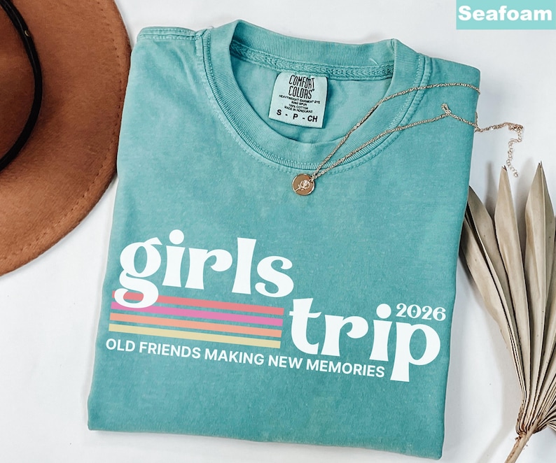 May include: Seafoam green t-shirt with the words "girls trip 2026" in white, with a colorful striped design. The shirt also has the text "OLD FRIENDS MAKING NEW MEMORIES". A gold necklace is draped across the shirt.