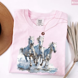 May include: A light pink Comfort Colors t-shirt featuring a watercolor image of three white horses running through water. A gold necklace with a pendant rests on the shirt. The shirt is folded, with the tag visible.