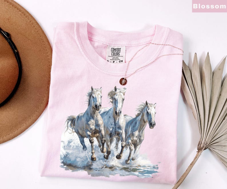 May include: A light pink Comfort Colors t-shirt featuring a watercolor image of three white horses running through water. A gold necklace with a pendant rests on the shirt. The shirt is folded, with the tag visible.