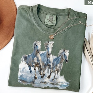 May include: Moss green t-shirt featuring a graphic of three white horses running through water. The shirt has a crew neck and short sleeves. A gold necklace with a pendant is draped across the shirt.