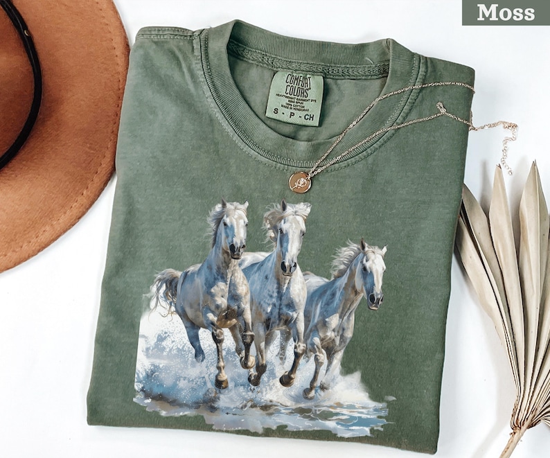 May include: Moss green t-shirt featuring a graphic of three white horses running through water. The shirt has a crew neck and short sleeves. A gold necklace with a pendant is draped across the shirt.