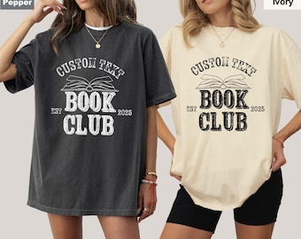 May include: Two women wearing oversized t-shirts with a vintage-style graphic design. The design features a book with the text "Custom Text Book Club Est 2025". The shirts are available in two colors: pepper and ivory.