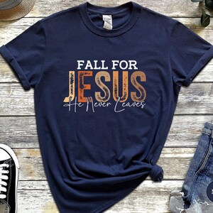 K&ouml;nnte beinhalten: Marineblaues T-Shirt mit dem Text "FALL FOR JESUS He Never Leaves" in Wei&szlig;, Orange und Braun. Das Shirt hat einen klassischen Rundhalsausschnitt.