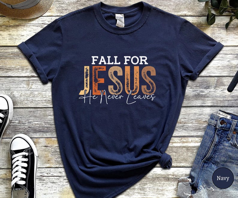 K&ouml;nnte beinhalten: Marineblaues T-Shirt mit dem Text "FALL FOR JESUS He Never Leaves" in Wei&szlig;, Orange und Braun. Das Shirt hat einen klassischen Rundhalsausschnitt.