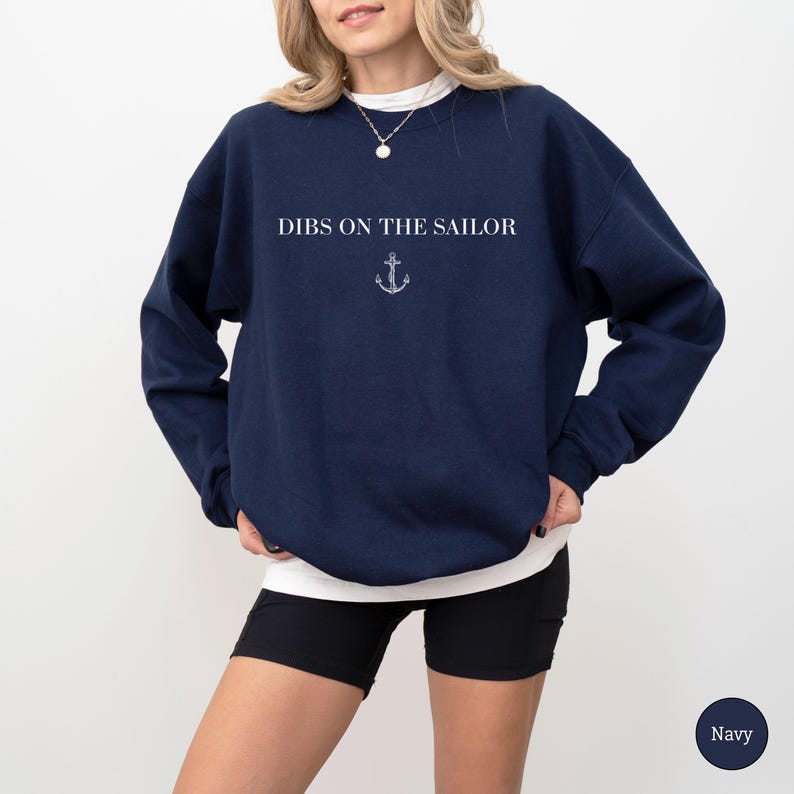 May include: Navy blue sweatshirt with the text "DIBS ON THE SAILOR" in white, and a small anchor graphic. The sweatshirt is a crew neck style. The model is wearing black shorts and a white undershirt.