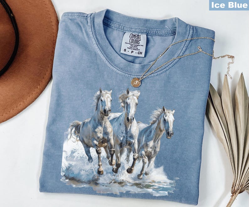 May include: Ice blue Comfort Colors t-shirt featuring a graphic of three white horses running through water. The shirt has a crew neck and short sleeves. A gold necklace with a pendant is draped across the shirt.