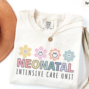 May include: Ivory-colored t-shirt with the words "NEONATAL INTENSIVE CARE UNIT" in colorful letters. Above the text are four smiling flower graphics in yellow, pink, orange, and blue. The shirt is made of a soft material.