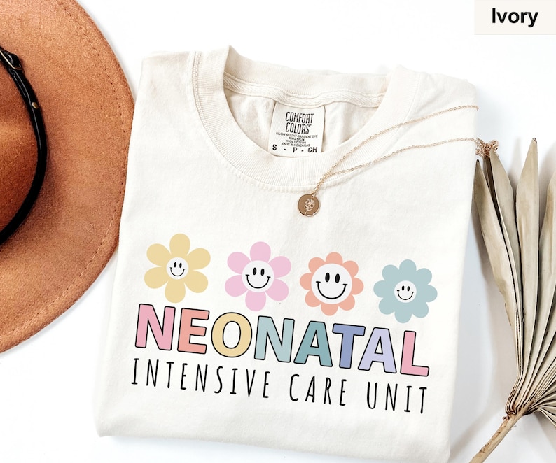 May include: Ivory-colored t-shirt with the words "NEONATAL INTENSIVE CARE UNIT" in colorful letters. Above the text are four smiling flower graphics in yellow, pink, orange, and blue. The shirt is made of a soft material.
