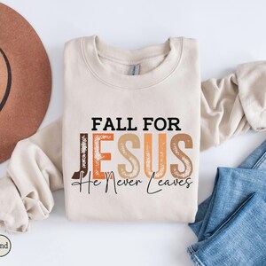 K&ouml;nnte beinhalten: Ein sandfarbenes Sweatshirt mit dem Text "FALL FOR JESUS He Never Leaves" in Schwarz, Braun und Orange. Das Sweatshirt liegt gefaltet auf einer wei&szlig;en Oberfl&auml;che neben einem braunen Hut und blauen Jeans.