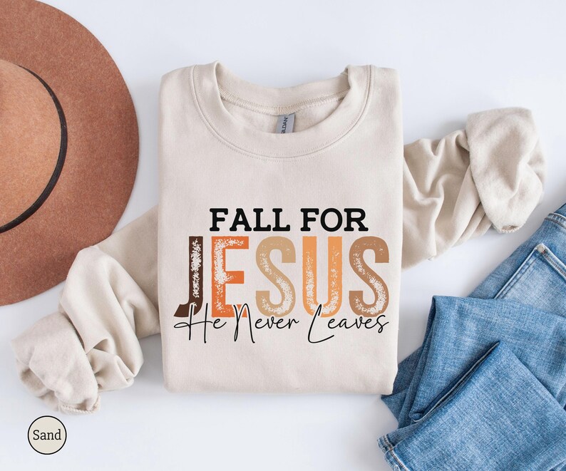 K&ouml;nnte beinhalten: Ein sandfarbenes Sweatshirt mit dem Text "FALL FOR JESUS He Never Leaves" in Schwarz, Braun und Orange. Das Sweatshirt liegt gefaltet auf einer wei&szlig;en Oberfl&auml;che neben einem braunen Hut und blauen Jeans.