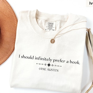 Op de afbeelding: Ivoor T-shirt met de quote "I should infinitely prefer a book." van Jane Austen. Het shirt heeft een ronde hals en korte mouwen. Een gouden ketting en een bruine hoed staan ook op de foto.