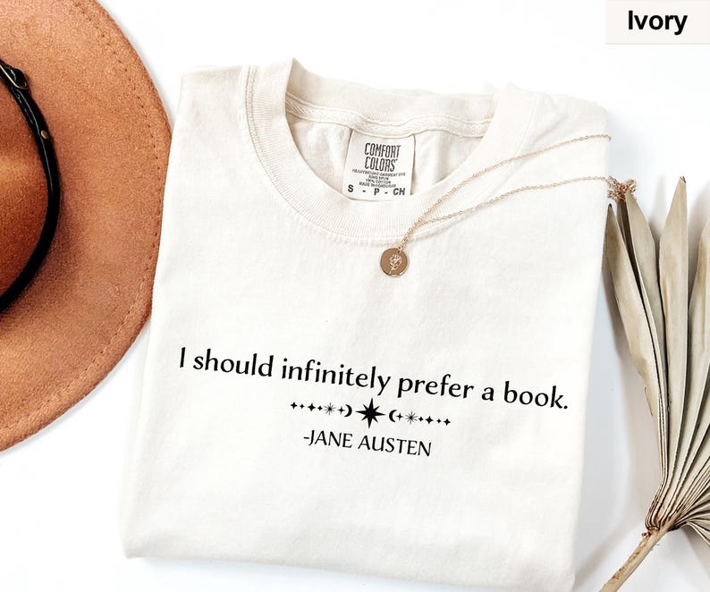 Op de afbeelding: Ivoor T-shirt met de quote "I should infinitely prefer a book." van Jane Austen. Het shirt heeft een ronde hals en korte mouwen. Een gouden ketting en een bruine hoed staan ook op de foto.