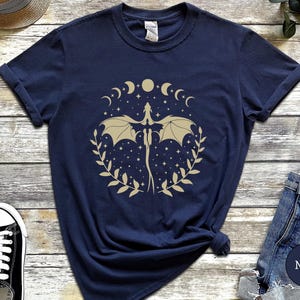 May include: Navy blue t-shirt with a gold graphic of a dragon flying through a crescent moon and stars. The dragon is surrounded by a wreath of leaves.