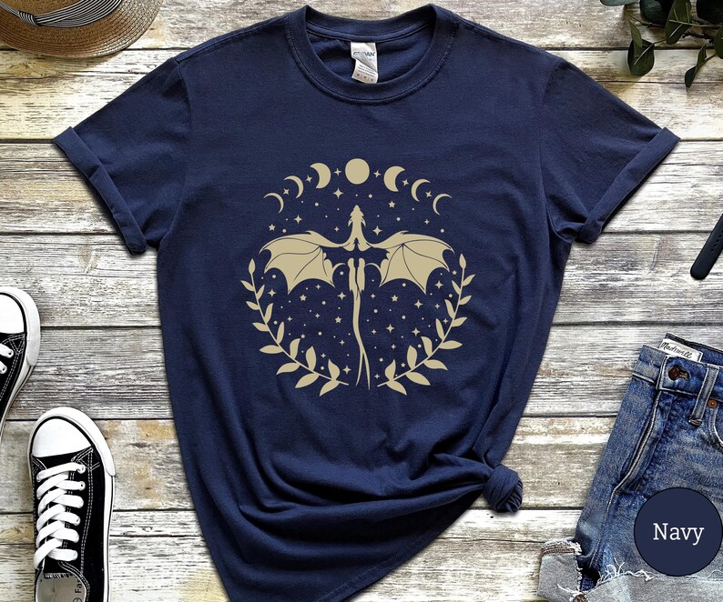 May include: Navy blue t-shirt with a gold graphic of a dragon flying through a crescent moon and stars. The dragon is surrounded by a wreath of leaves.
