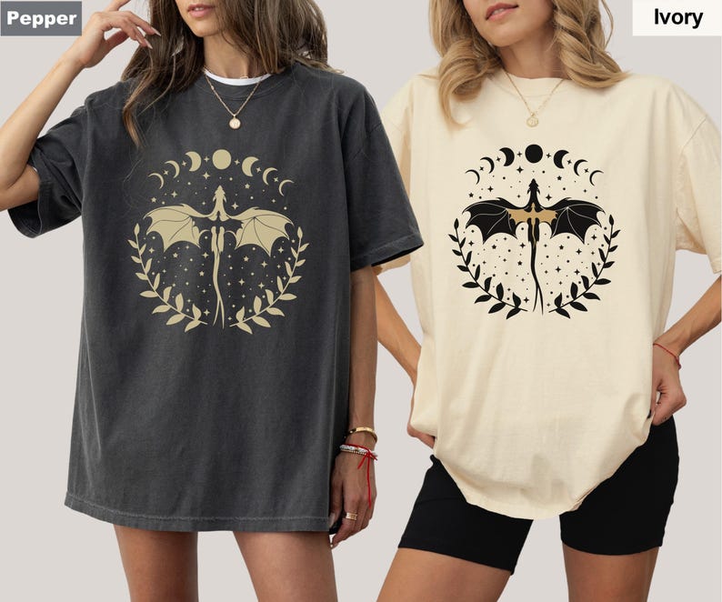 May include: Two women wearing oversized t-shirts with a graphic print of a dragon with wings spread, surrounded by a circle of moons and stars. The t-shirt on the left is dark gray and the t-shirt on the right is ivory.