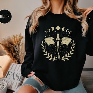 May include: Black sweatshirt with a graphic print of a dragon with wings spread, surrounded by a circle of crescent moons and stars. The text "Black" is visible in the top left corner of the image.