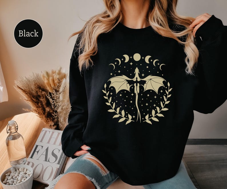 May include: Black sweatshirt with a graphic print of a dragon with wings spread, surrounded by a circle of crescent moons and stars. The text "Black" is visible in the top left corner of the image.