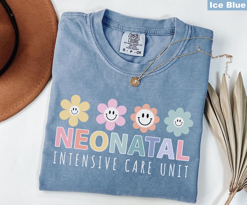 May include: Ice blue t-shirt with a floral design and the words "Neonatal Intensive Care Unit" in pastel colors. The shirt has a crew neck and short sleeves. A gold necklace with a pendant is draped across the shirt.