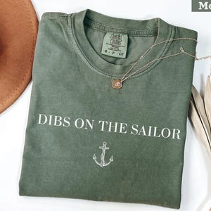 May include: Moss green t-shirt with the phrase "Dibs on the Sailor" in white block letters and an anchor graphic. A gold-colored necklace with a pendant rests on the shirt. The shirt is folded and displayed with a brown hat and dried plant.
