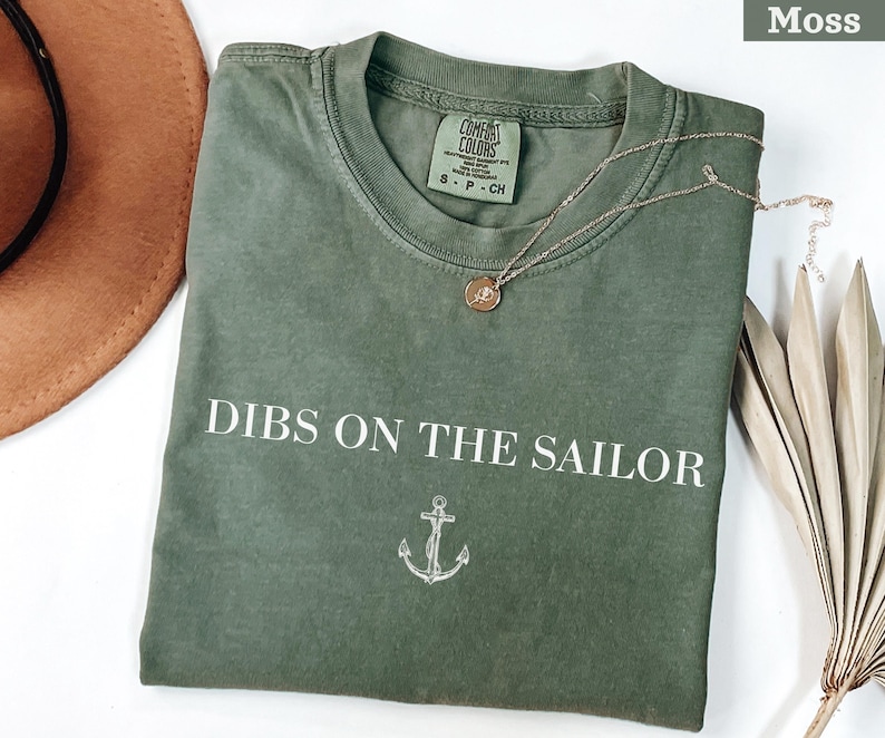 May include: Moss green t-shirt with the phrase "Dibs on the Sailor" in white block letters and an anchor graphic. A gold-colored necklace with a pendant rests on the shirt. The shirt is folded and displayed with a brown hat and dried plant.