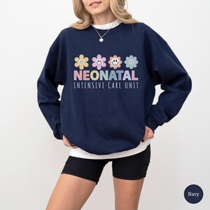 May include: Navy blue sweatshirt with the text "NEONATAL INTENSIVE CARE UNIT" in white. Above the text are four smiling flower graphics in pastel colors. The sweatshirt is a crew neck style.