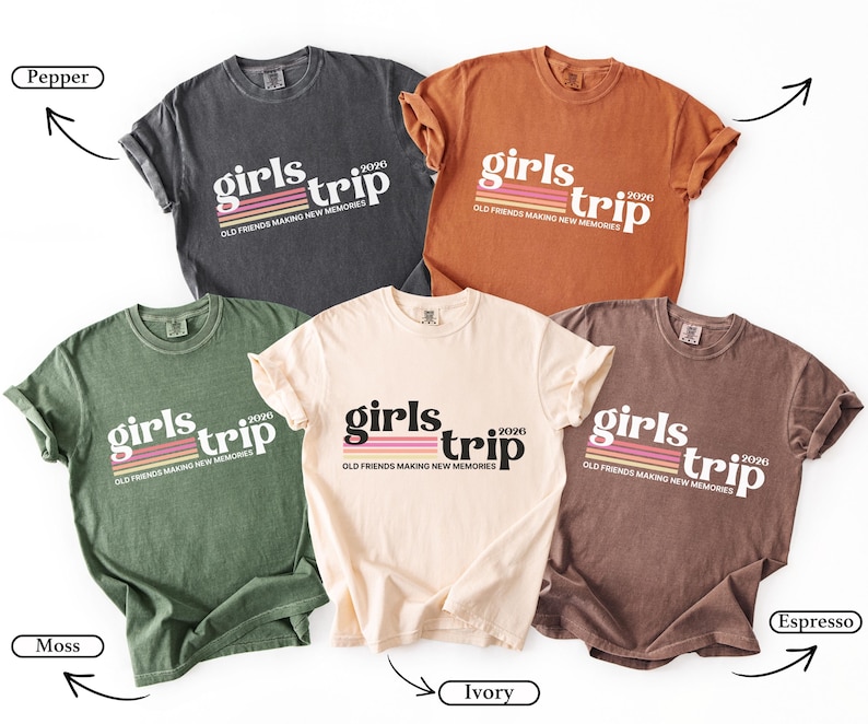 May include: Five different colored t-shirts with the text "girls trip 2026" in a retro font. The shirts are in the colors pepper, moss, ivory, espresso, and a burnt orange. The text "Old friends making new memories" is below the main text.