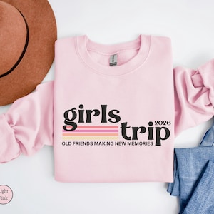 May include: A light pink sweatshirt with the words "girls trip 2026" in black and a striped design. The phrase "OLD FRIENDS MAKING NEW MEMORIES" is below the text. A brown hat and blue jeans are also in the image.