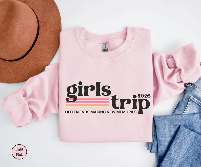 May include: A light pink sweatshirt with the words "girls trip 2026" in black and a striped design. The phrase "OLD FRIENDS MAKING NEW MEMORIES" is below the text. A brown hat and blue jeans are also in the image.