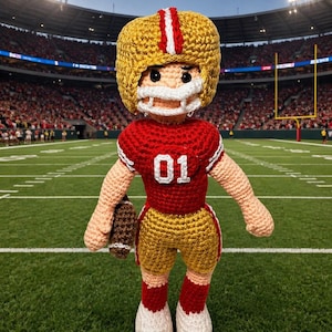 May include: A handmade crochet football player doll wearing a gold helmet with a red and white stripe, a red jersey with the number 01, and gold shorts. The doll is holding a brown football and is standing on a green field.