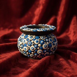 Peut inclure: Petit pot rond en céramique peint à la main, avec un bord et une base noirs. Le pot est décoré de motifs circulaires bleus et blancs. Le fond est un tissu rouge foncé texturé.