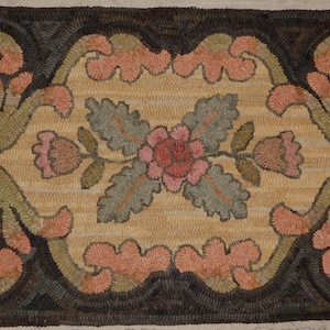 May include: Hand-hooked rug with a central floral design. The rug features a tan background with a dark brown border. The floral design includes pink, green, and tan elements, creating a decorative and textured appearance. The rug is rectangular.